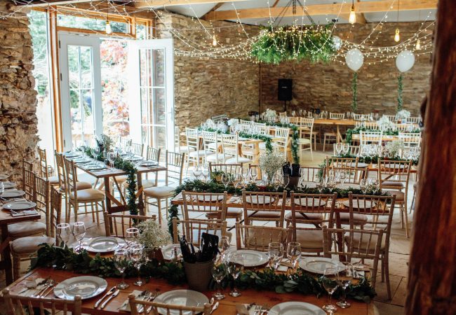 Inside of the Stone Barn with tables and chairs set for the wedding breakfast, fairy lights and greenery adorn the barn